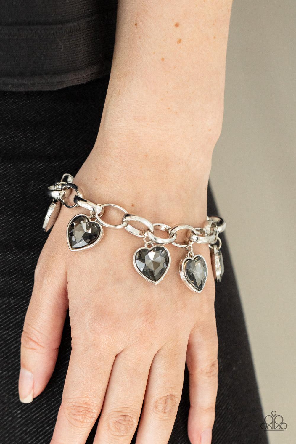 Paparazzi Accessories Candy Heart Charmer - Silver Smoky heart-shaped gems are encased in sleek silver frames that swing from an oversized silver chain, creating a sparkly fringe around the wrist. Features an adjustable clasp closure. Sold as one individu