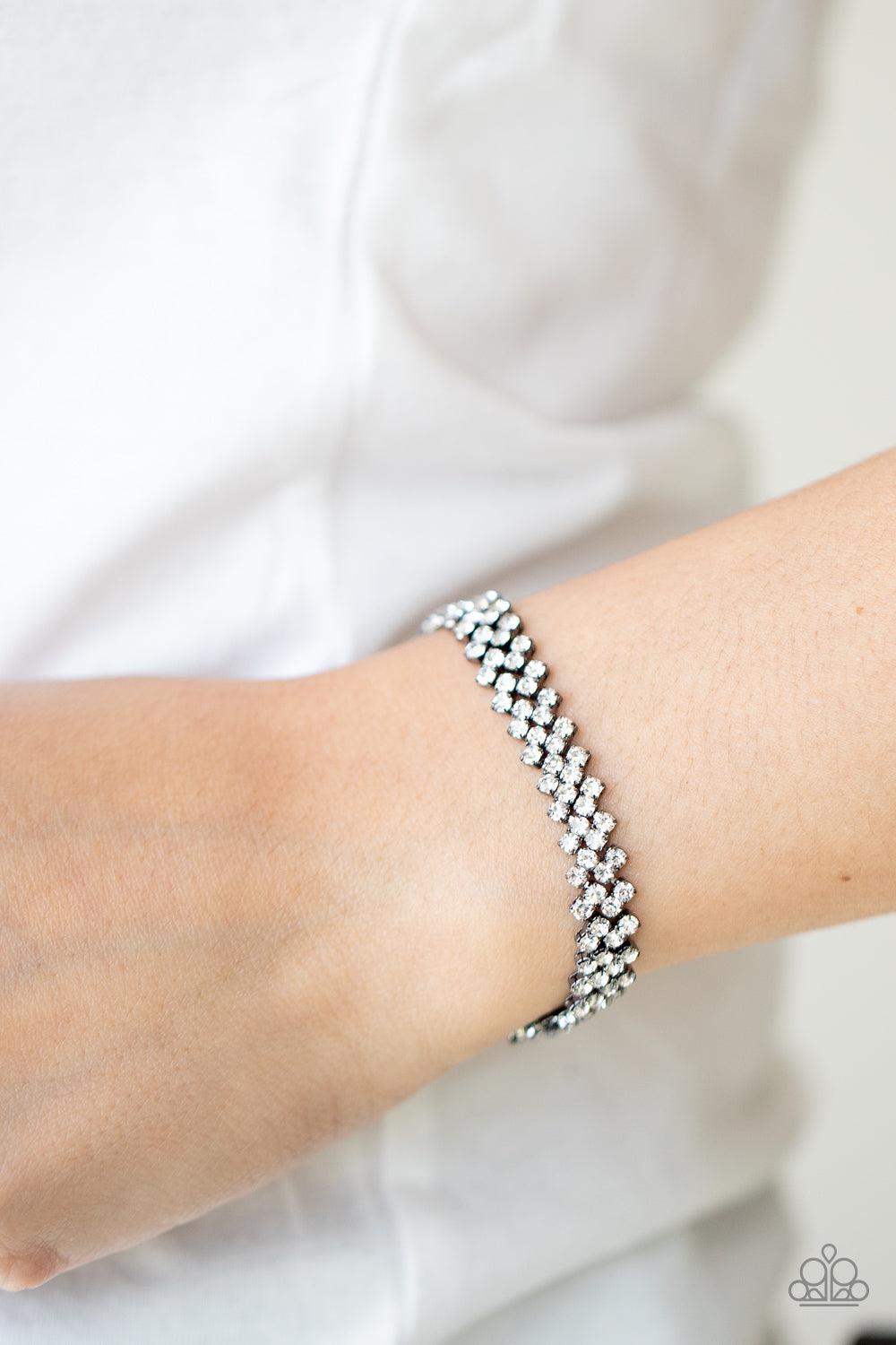 Paparazzi Accessories Chicly Candescent - Black Featuring sleek gunmetal fittings, dainty rows of glassy white rhinestones delicately slant across the wrist, coalescing into a timeless centerpiece. Features an adjustable clasp closure. Sold as one individ