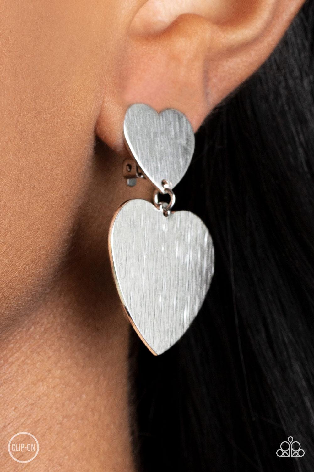 Paparazzi Accessories Cowgirl Crush - Silver *Clip-On Featuring a shimmery scratched finish, an oversized silver heart charm swings from the bottom of a smaller silver heart charm, resulting in a flirtatious lure. Earring attaches to a standard clip-on fi