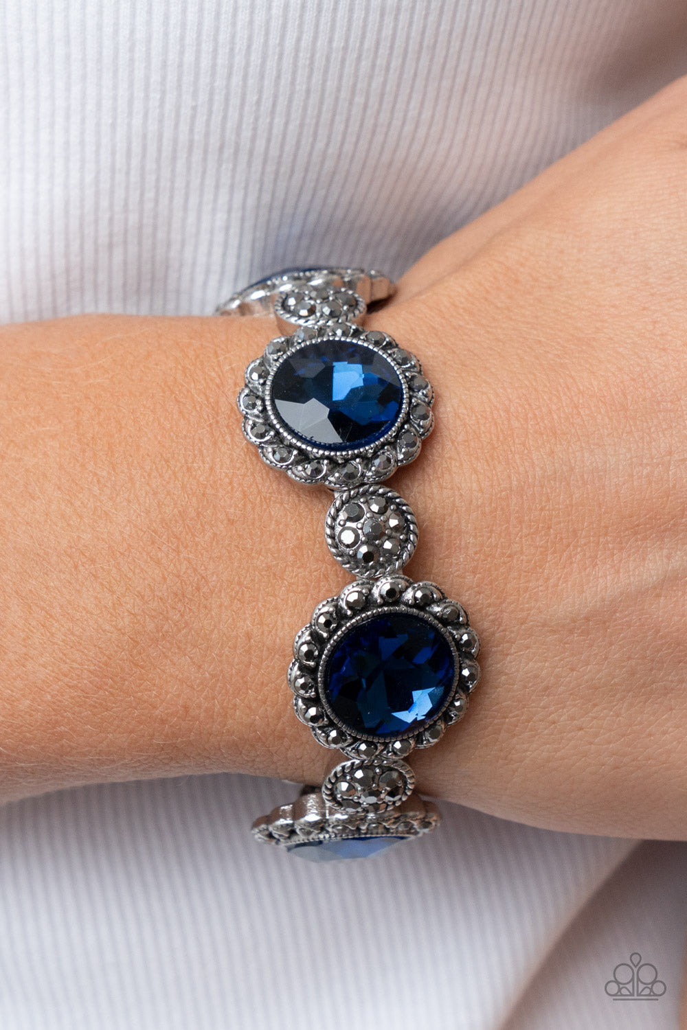 Paparazzi Accessories Palace Property - Blue An oversized blue rhinestone adorns the center of a hematite rhinestone petaled floral frame. Infused with hematite dotted silver accents, the glitzy floral frames sparkle along stretchy bands around the wrist
