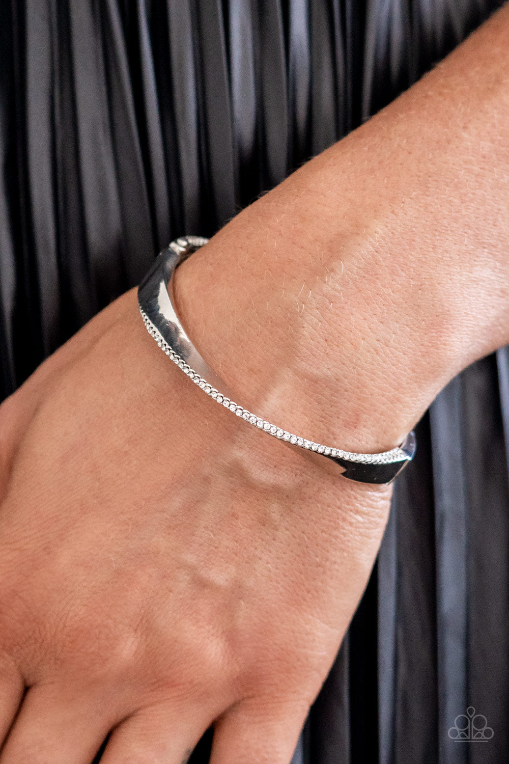 Paparazzi Accessories Artistically Adorned - White An oversized, warped, shiny silver cuff wraps around the wrist in an artistic manner. A dainty row of white rhinestones encrusts along the curves of the cuff for an understated, elegant dazzle. Features a