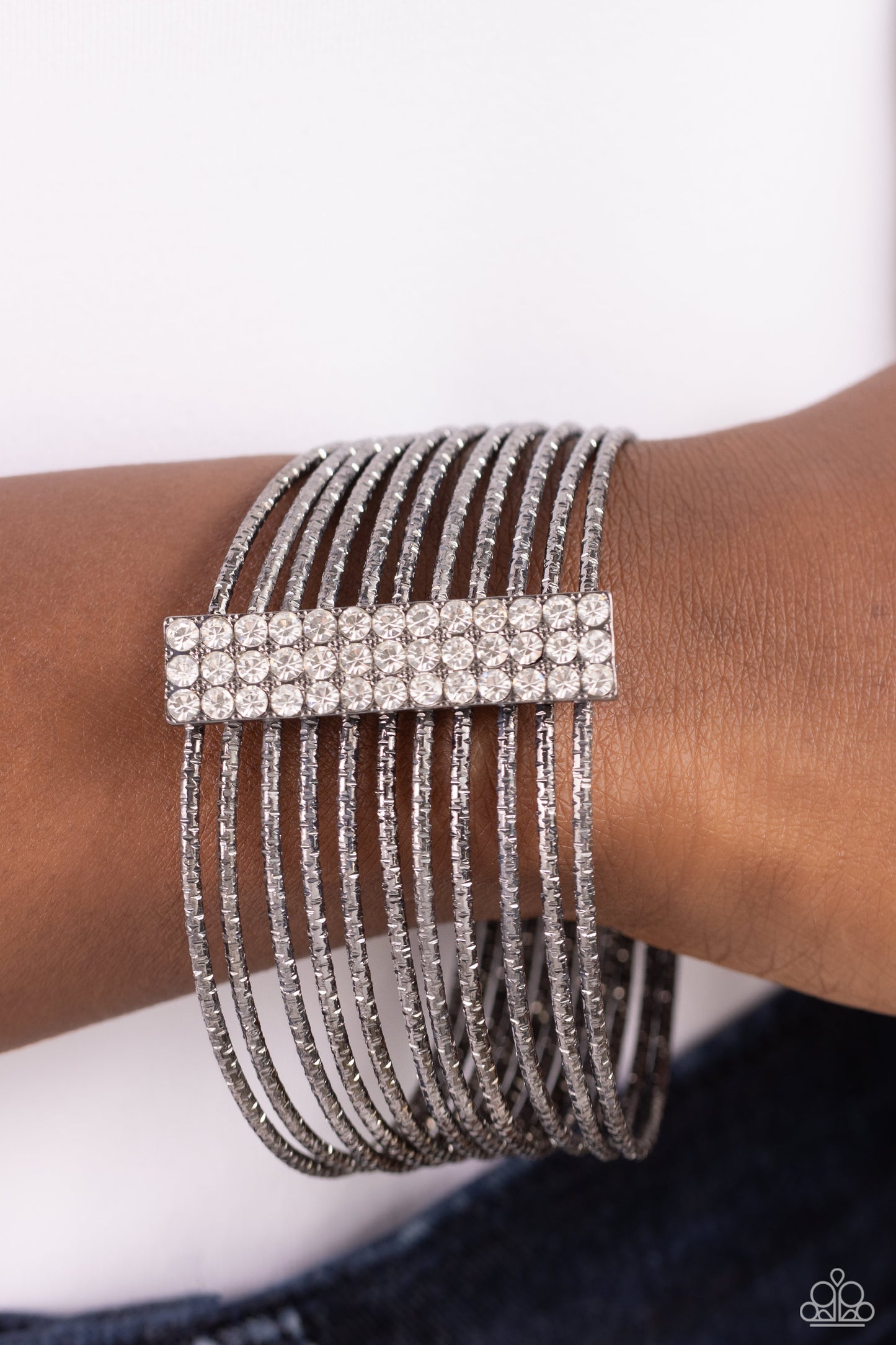 Paparazzi Accessories Shimmery Silhouette - Black Held together by an elongated rectangular gunmetal fitting and encrusted in three rows of white gems, gunmetal bangles embossed in shimmery diamond-cut textures stack across the wrist for a refined display