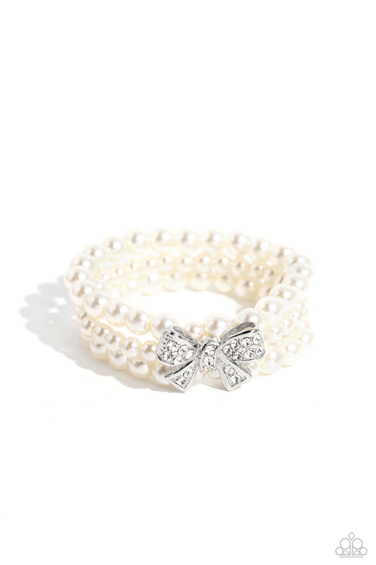 Paparazzi Accessories How Do You Do? - White Strung along elastic stretchy bands, three strands of high-sheen classic white pearls wrap around the wrist. Featured atop the pearly collection, a loopy silver bow, filled with glistening white rhinestones is