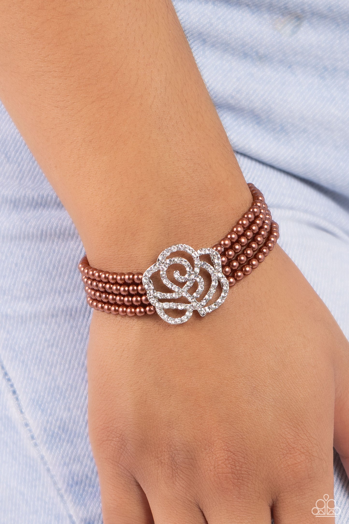 Paparazzi Accessories Regal Rose - Brown Held together by dainty silver plates, four strands of classic brown pearls are threaded around the wrist. Encrusted in glassy white rhinestones, a glitzy silver rose blooms at the center of the wrist for a fierce