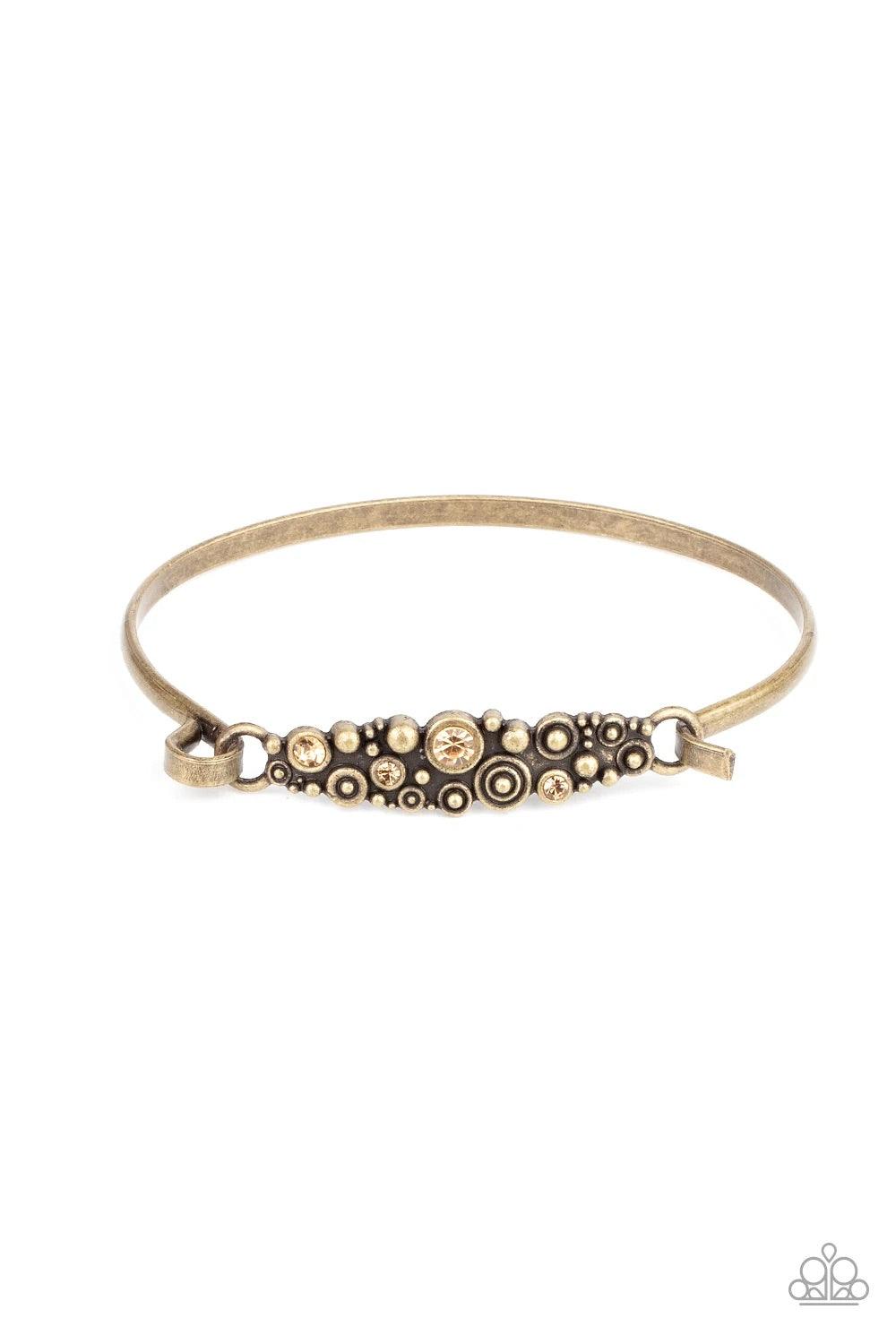 Paparazzi Accessories Bubbling Whimsy - Brass A cluster of engraved and antiqued brass swirls merge with topaz rhinestones and coalesce into a whimsical display across the wrist on a dainty brass cuff. Features a hook and eye closure. Sold as one individu