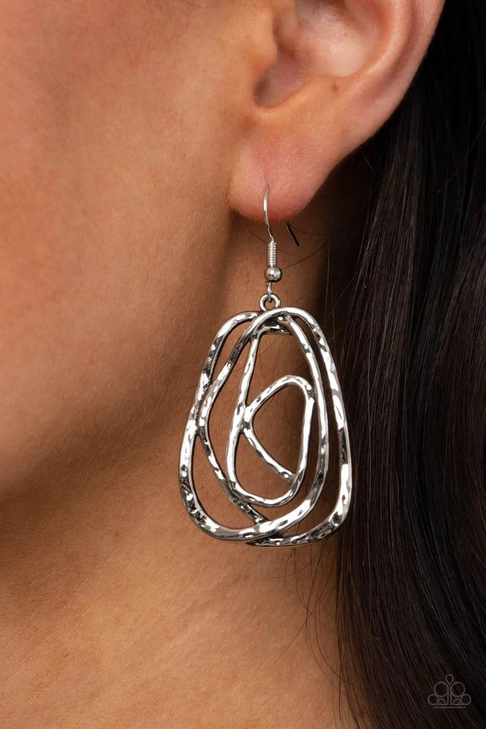 Paparazzi Accessories Artisan Relic - Silver Featuring a hammered finish, a rustic silver wire delicately wraps into an asymmetrical frame for a dizzying artisan inspired look. Earring attaches to a standard fishhook fitting. Sold as one pair of earrings.
