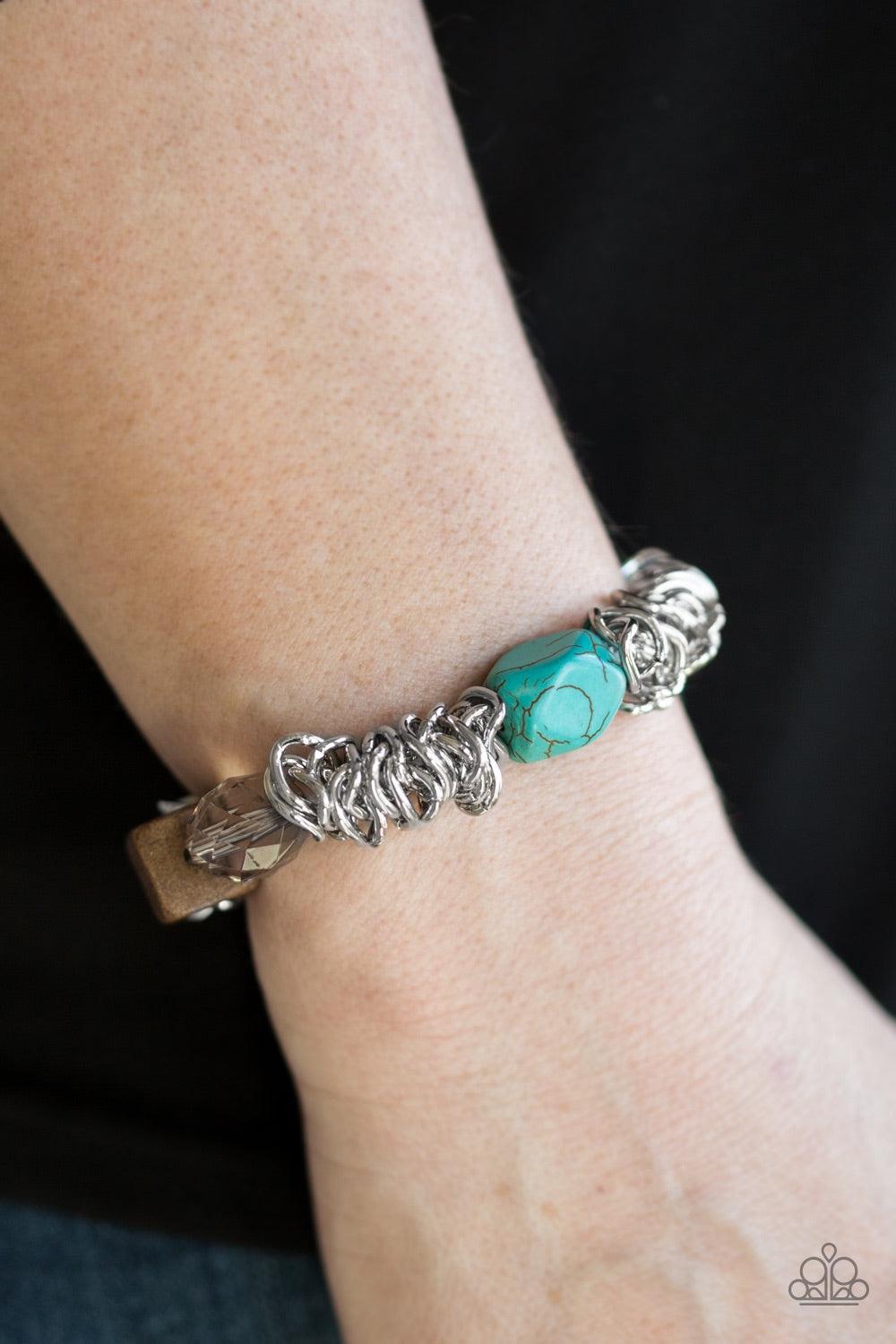Paparazzi Accessories Mesmerizingly Magmatic - Blue A mishmash of bold silver chain, refreshing turquoise stones, a square wooden bead and metallic and crystal-like accents are threaded along a stretchy elastic band for a seasonal look. Jewelry