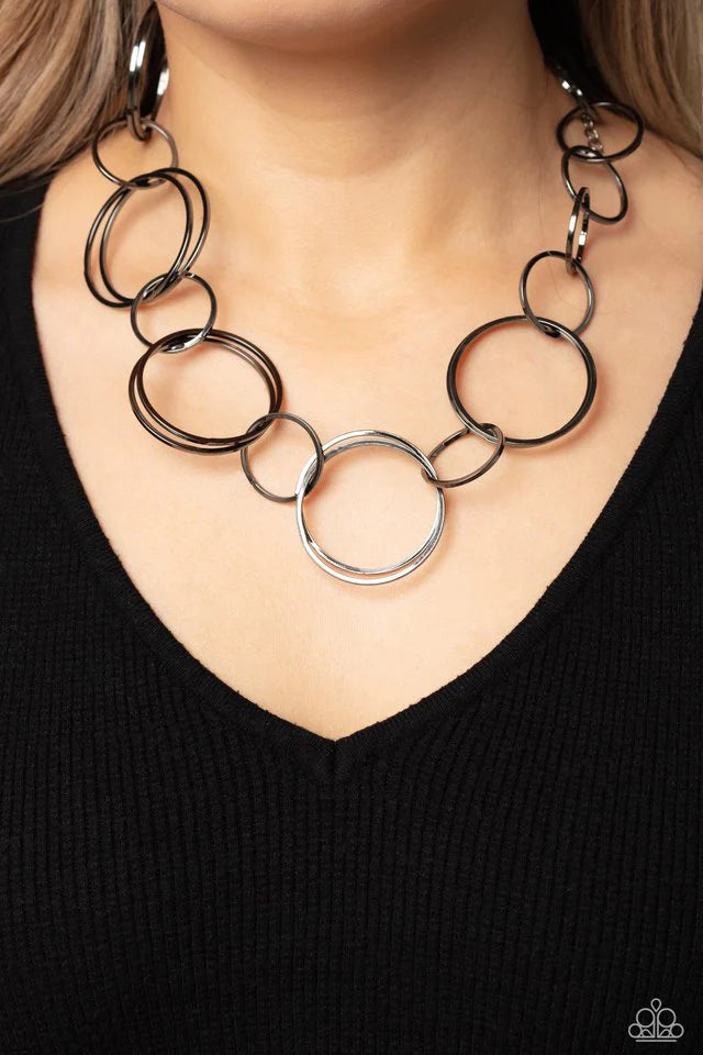 Paparazzi Accessories Shimmering Symphony - Black A shimmering collection of thin gunmetal hoops in varying sizes interlock and coalesce down the neckline for a refined monochromatic statement piece. Features an adjustable clasp closure. Sold as one indiv