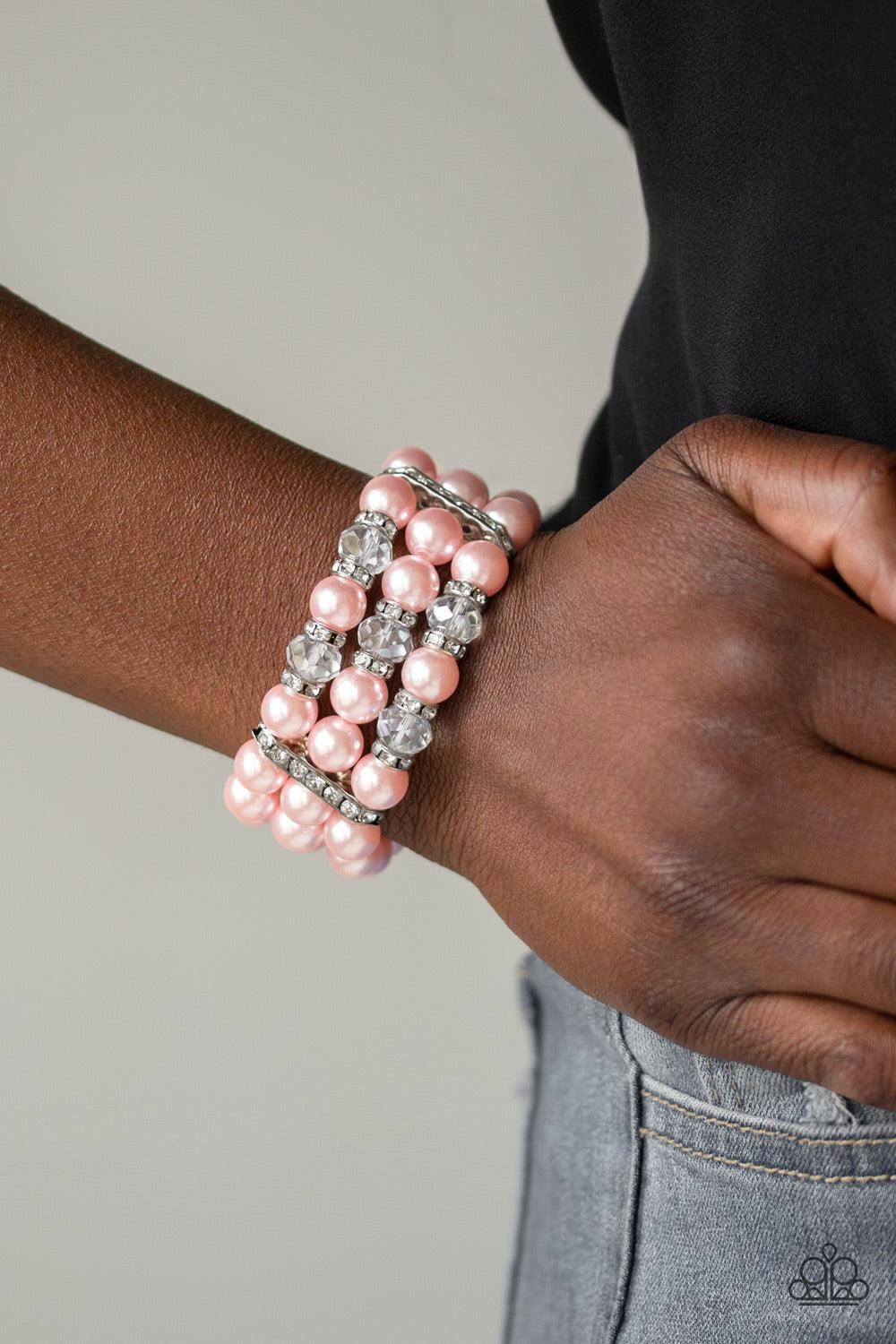 Paparazzi Accessories Undeniably Dapper - Pink Pinched between white rhinestone encrusted frames, white rhinestone encrusted rings, crystal-beads, and pink pearls are threaded along elastic stretchy bands for a glamorous look. Sold as one individual brace