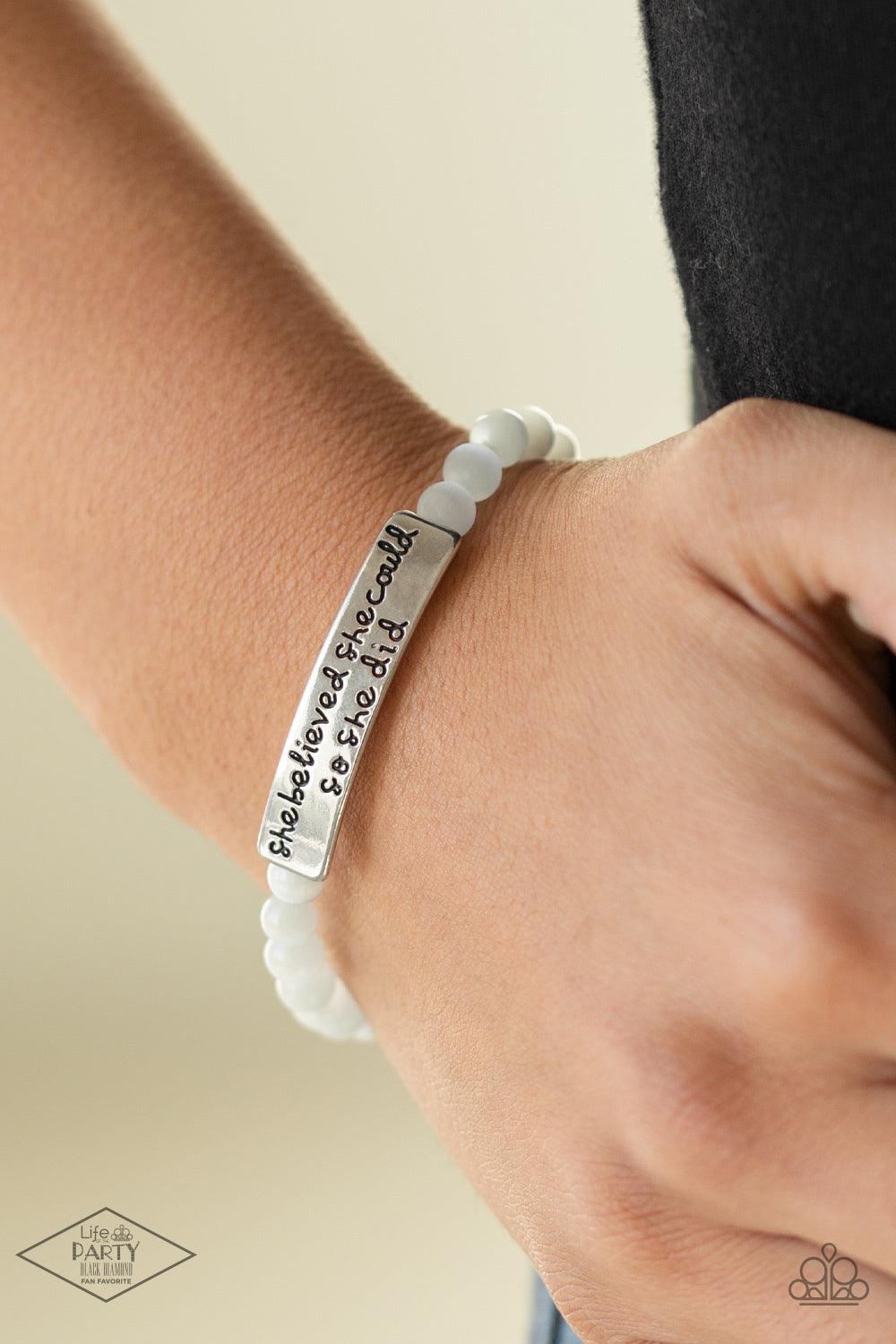 Paparazzi Accessories So She Did - White A collection of dainty white cat’s eye stone beads and an antiqued frame stamped in the inspirational phrase, “She believed she could so she did” are threaded along a stretchy band around the wrist for a whimsical