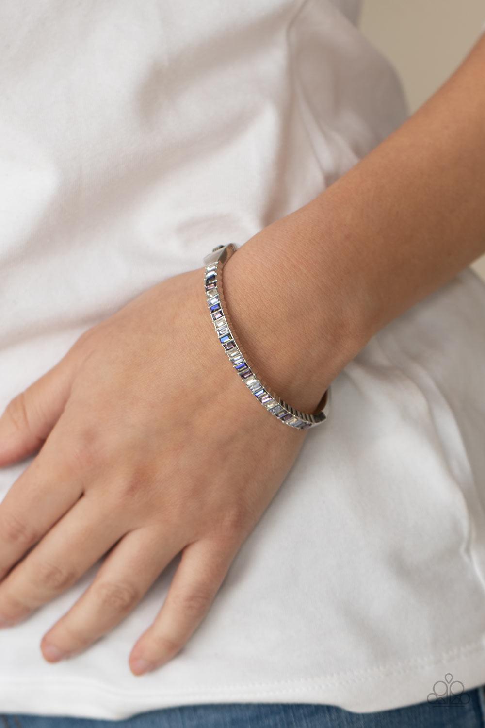 Paparazzi Accessories Toast to Twinkle - Purple Featuring regal emerald style cuts, a dainty row of purple, white, and blue rhinestones are encrusted across the front of a silver cuff-like bangle for a timeless fashion. Features a hinged closure. Sold as