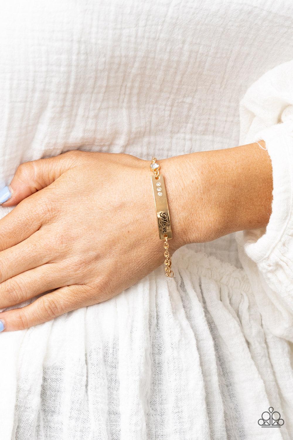 Paparazzi Accessories Mom Always Knows - Gold Infused with a glittery white rhinestone charm, a curved gold plate is dotted in dainty white rhinestones and stamped in the word, "Mom," as it delicately attaches to a dainty gold chain around the wrist for a
