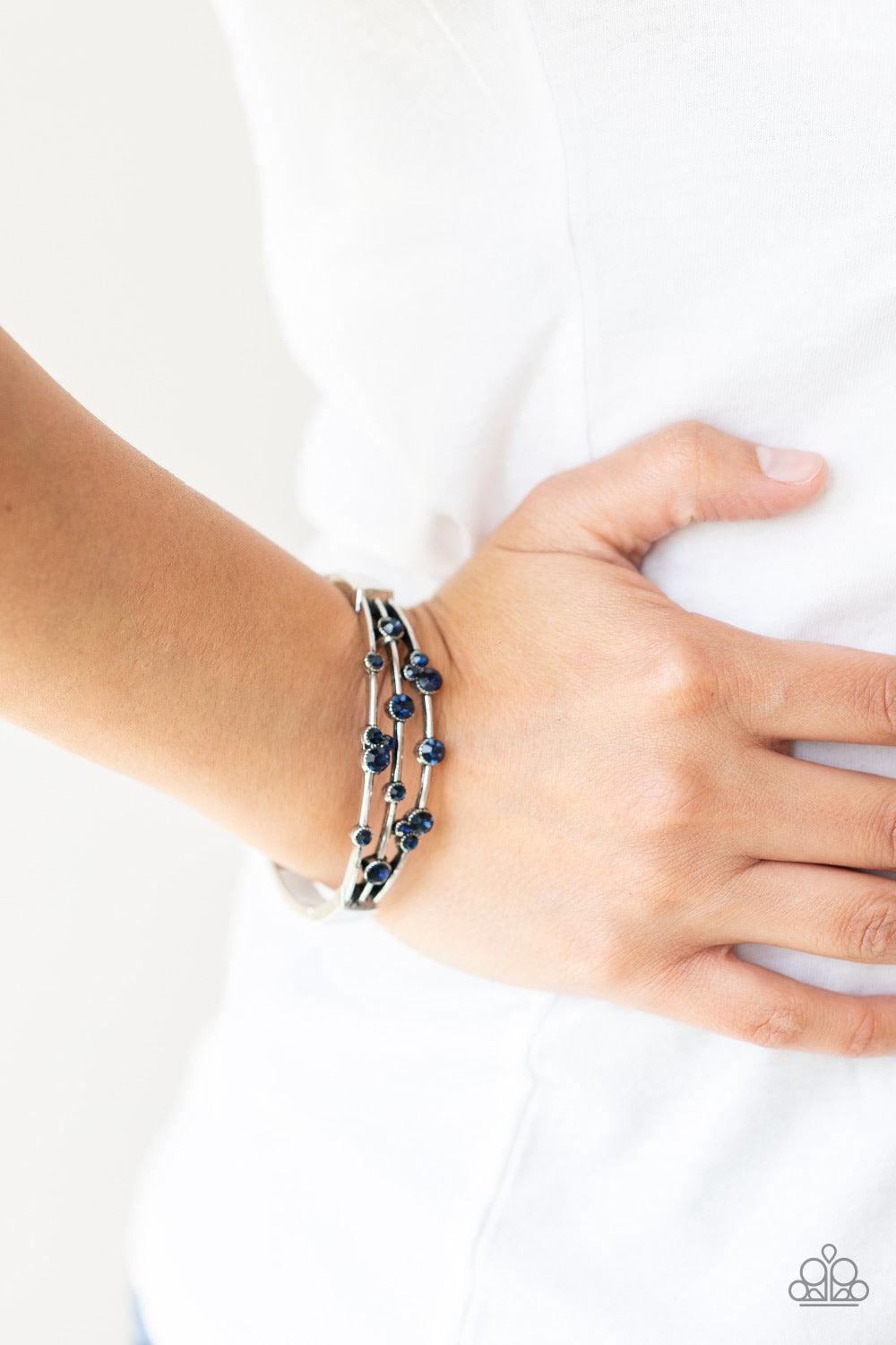 Paparazzi Accessories Cosmic Candescence - Blue A smattering of glittery blue rhinestones adorn three silver bars that coalesce into a versatile silver cuff-like bangle around the wrist. Features a hinged closure. Sold as one individual bracelet. Jewelry