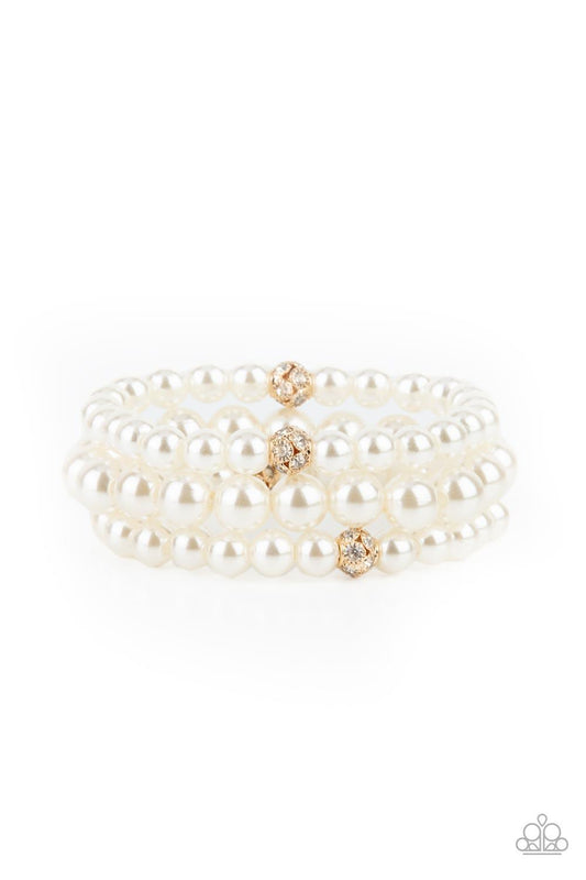 Paparazzi Accessories Here Comes The Heiress - Gold Infused with white rhinestone encrusted gold beads, a bubbly collection of mismatched white pearls are threaded along stretchy bands around the wrist for a vintage inspired layered look. Sold as one set