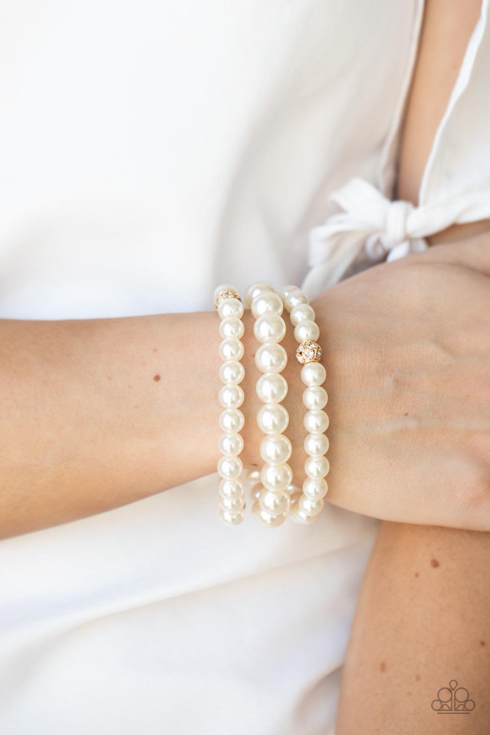 Paparazzi Accessories Here Comes The Heiress - Gold Infused with white rhinestone encrusted gold beads, a bubbly collection of mismatched white pearls are threaded along stretchy bands around the wrist for a vintage inspired layered look. Sold as one set