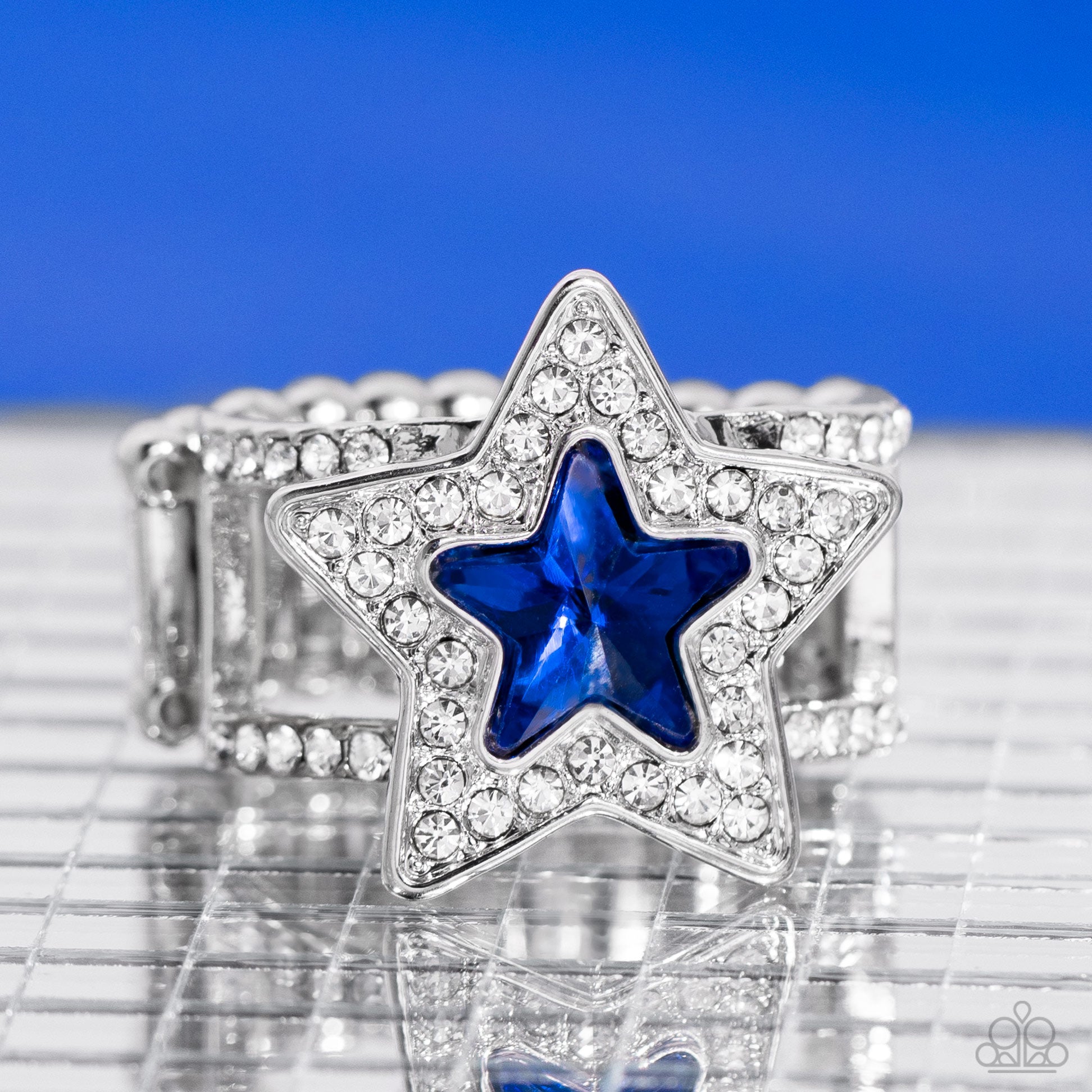 Paparazzi Accessories One Nation Under Sparkle - Blue A star shaped blue gem is pressed into the center of a silver star dotted in blinding white rhinestones, creating a stellar centerpiece atop two white rhinestone encrusted silver bands. Features a stre