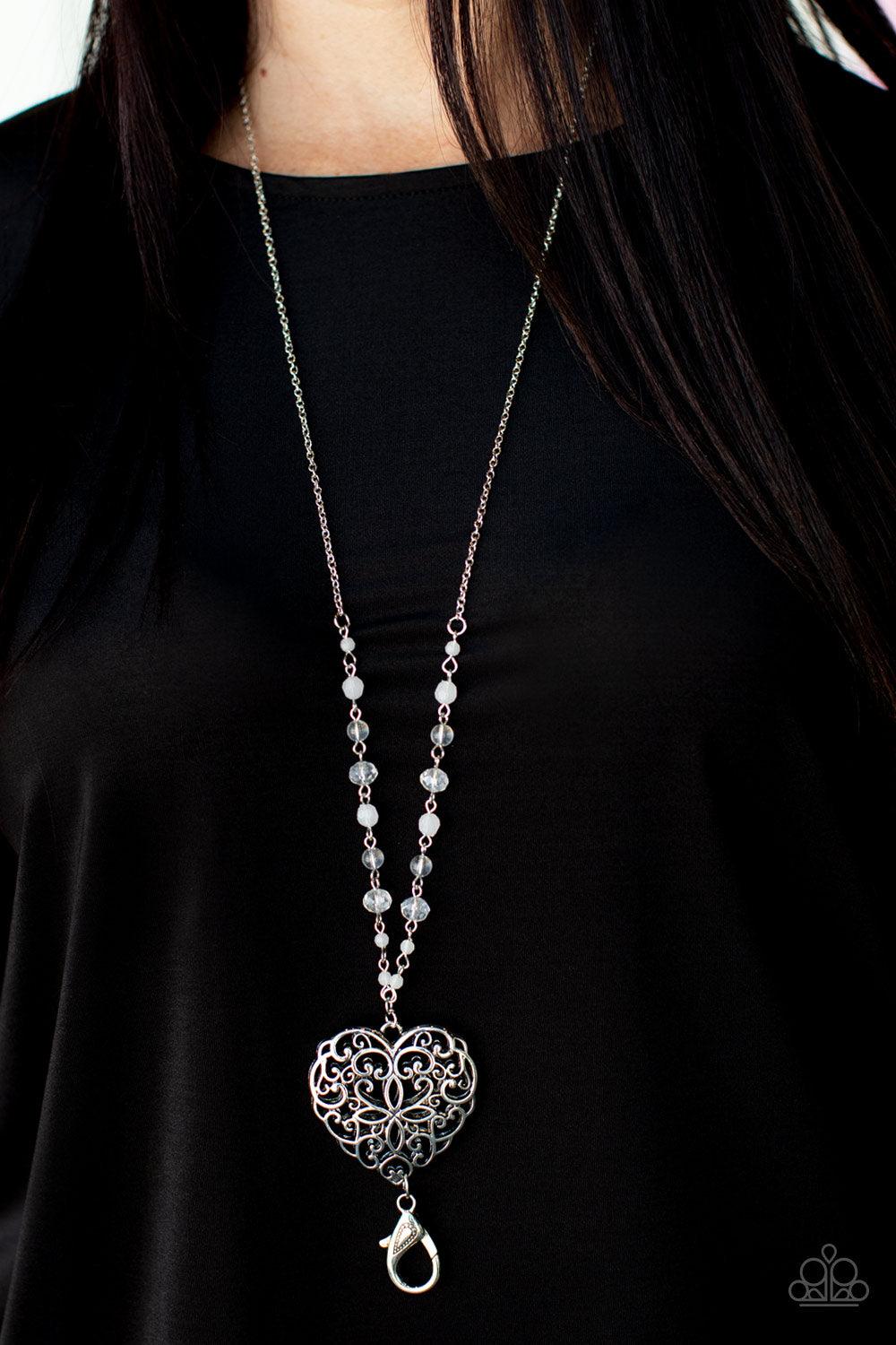 Paparazzi Accessories Doting Devotion - White *Lanyard A dainty collection of opaque, glassy, and crystal-like beads give way to an oversized filigree filled heart frame, creating an antiqued locket inspired pendant at the bottom of a lengthened silver ch