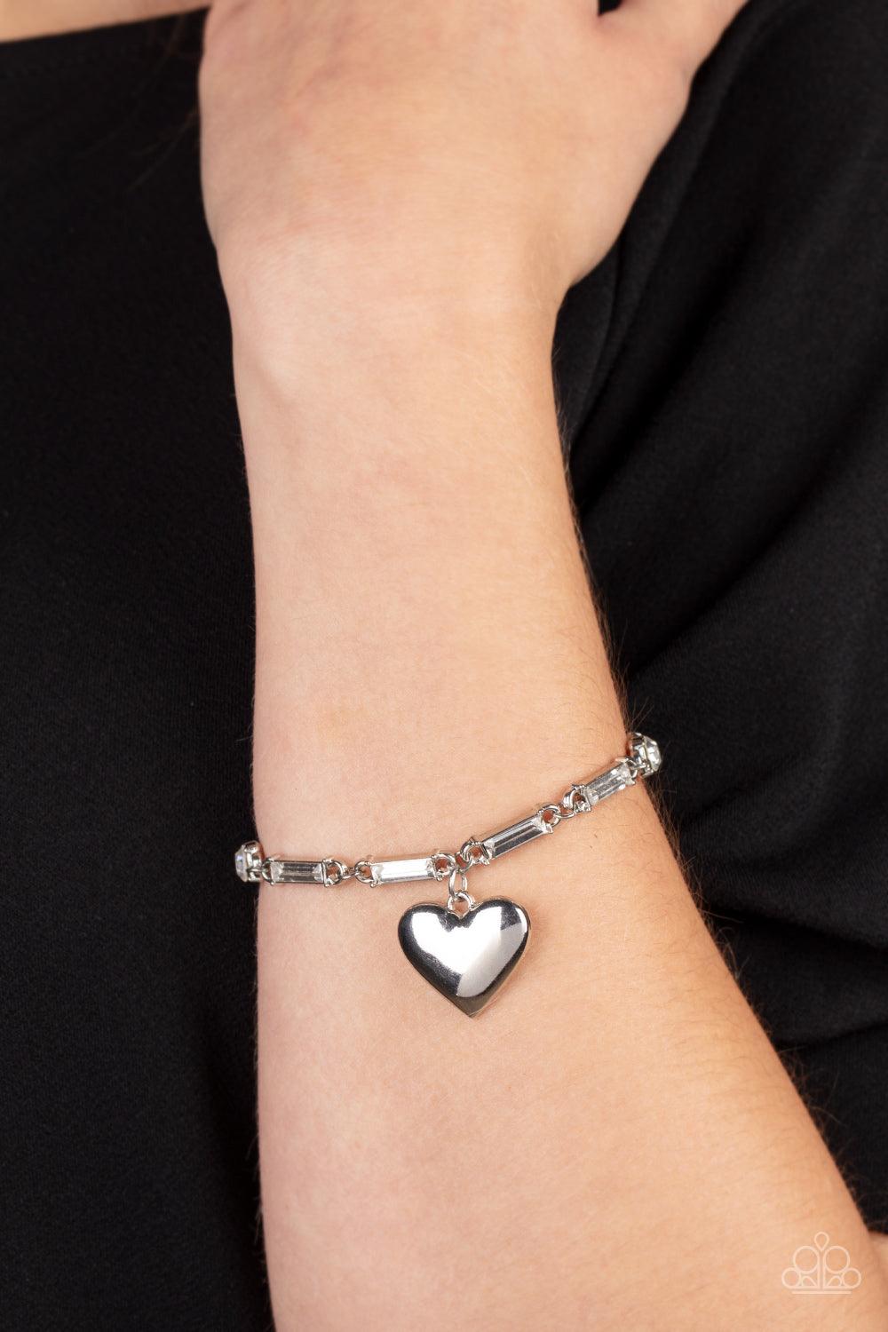Paparazzi Accessories Sweetheart Secrets - White Encased in sleek silver fittings, round and emerald cut rhinestones delicately link into a sparkly chain around the wrist. A shiny silver heart charm swings from the glittery compilation, adding a flirtatio