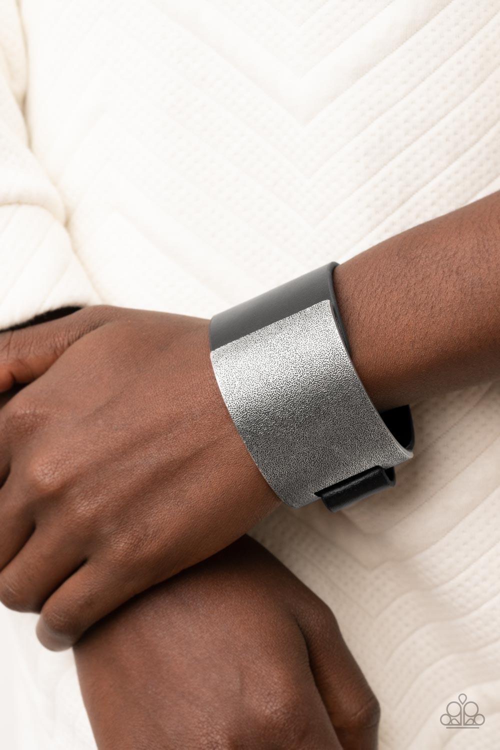 Paparazzi Accessories Studded Synchronism - Black A curved tin finished silver panel, coated in antiqued texture, attaches to a wide black leather band with silver snaps resulting in an edgy industrial element around the wrist. Features an adjustable snap