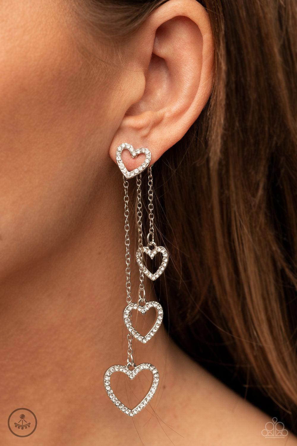 Paparazzi Accessories Falling In Love - White Encrusted in dainty white rhinestones, a glittery silver heart fitting attaches to a double-sided post designed to fasten behind the ear. Attached to dainty silver chains of varying lengths, a glittery collect