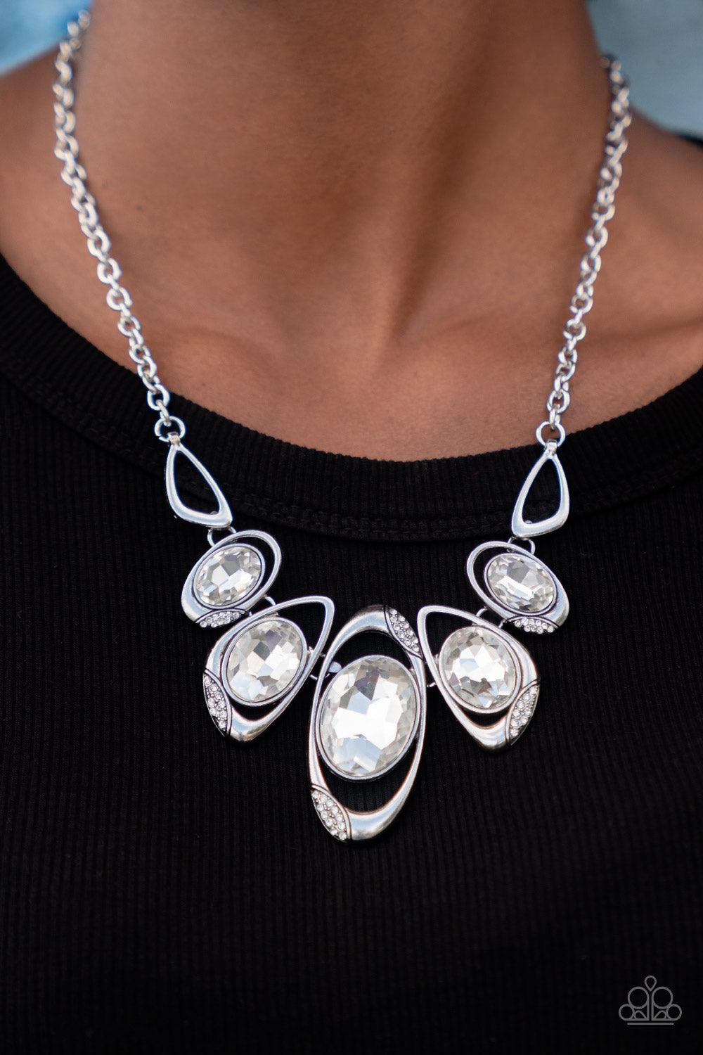 Paparazzi Accessories Hypnotic Twinkle - White Dusted in sections of glassy white rhinestones, asymmetrical silver frames curl around oversized white gems below the collar. Varying in shape, the mismatched frames increase in size as they near the center f