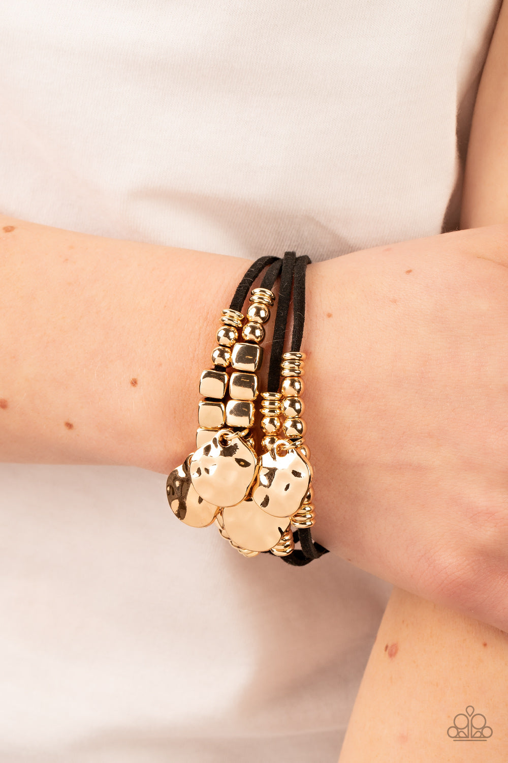 Paparazzi Accessories Token Trek - Gold Asymmetrical, hammered gold discs swing from layers of black suede that are adorned with gold discs, beads, and cubes, resulting in noisemaking shimmer around the wrist. Features an adjustable clasp closure. Sold as