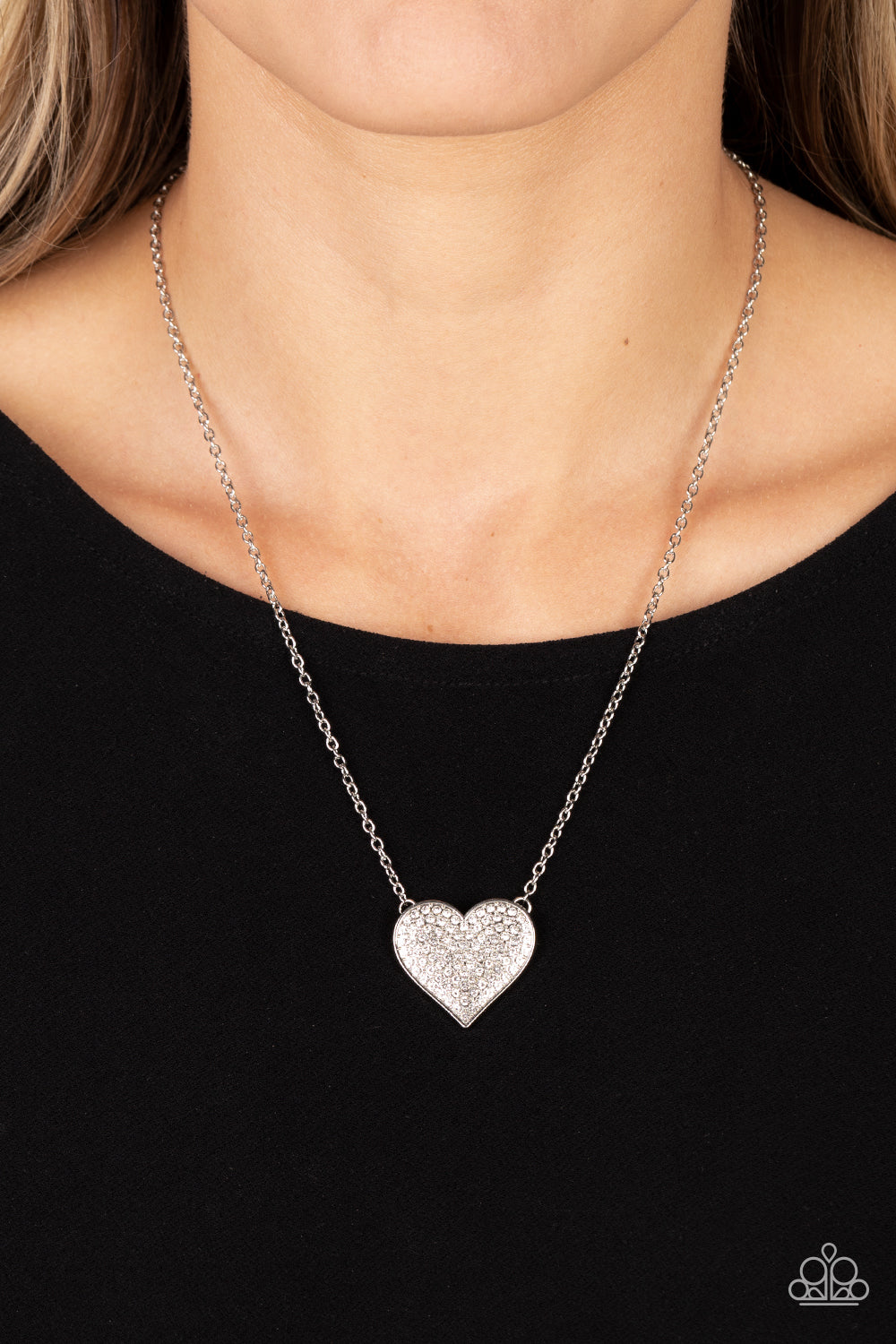 Paparazzi Accessories Spellbinding Sweetheart - White A heart-shaped pendant in a silver finish is anchored between two strands of delicate chain in the same reflective finish. The interior of the heart is filled with glittery white rhinestones, inviting