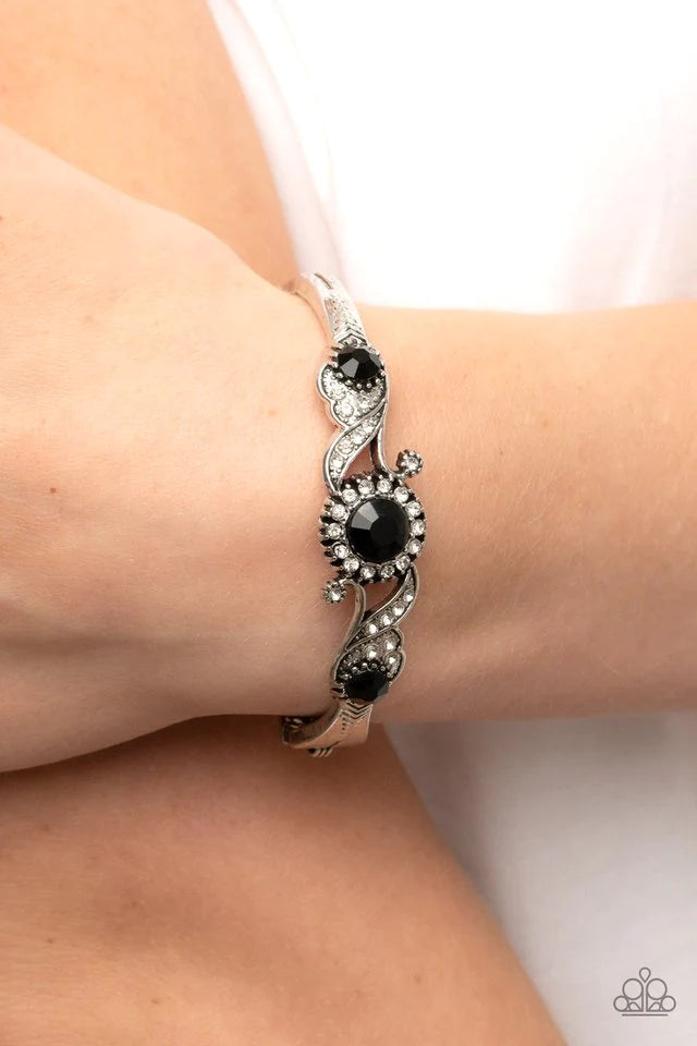 Paparazzi Accessories Expert Elegance - Black Dotted in dainty white rhinestones, leafy and floral silver frames swoop around black rhinestone centers atop a dainty silver bangle-like bracelet, resulting in an elegant centerpiece. Sold as one individual b