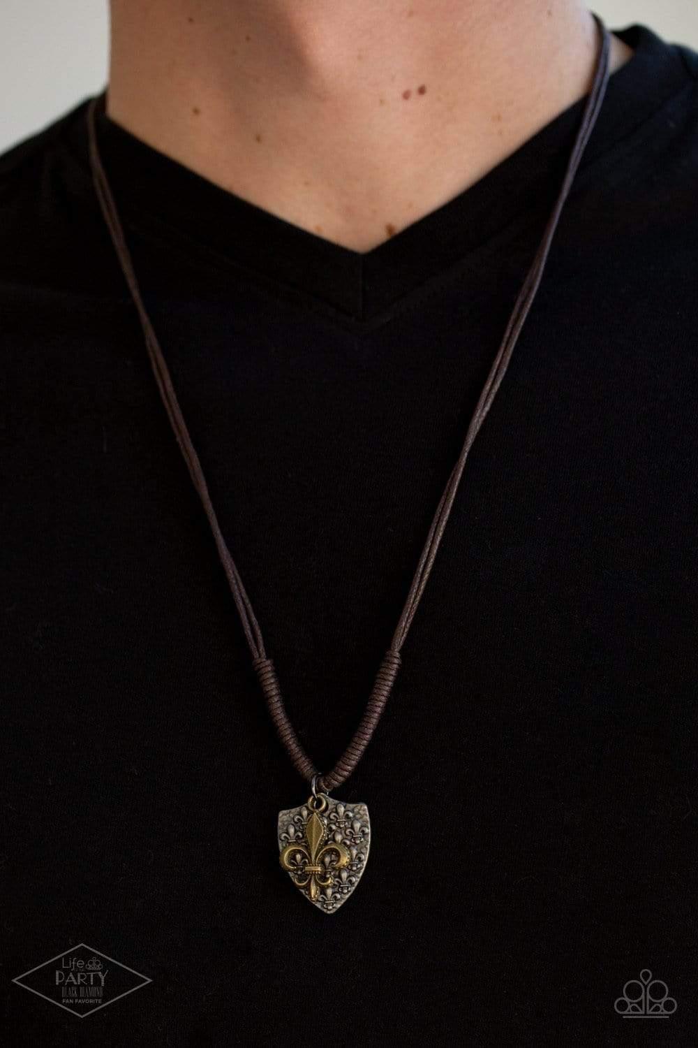 Paparazzi Accessories Shielded Simplicity - Brass An antiqued silver shield-shaped pendant embossed with a repeating fleur-de-lis pattern is paired with a brass fleur-de-lis charm at the bottom of doubled strands of brown cording. Sliding knots allow for