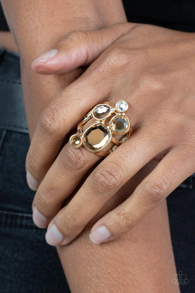 Paparazzi Accessories Demandingly Duchess - Brown A collection of sparkling champagne-colored gems in varying sizes, accented by a single white rhinestone, forms an asymmetrical cluster atop the finger. Set in sleek gold frames over airy gold bands, the b