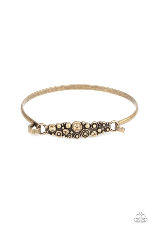 Paparazzi Accessories Bubbling Whimsy - Brass A cluster of engraved and antiqued brass swirls merge with topaz rhinestones and coalesce into a whimsical display across the wrist on a dainty brass cuff. Features a hook and eye closure. Sold as one individu