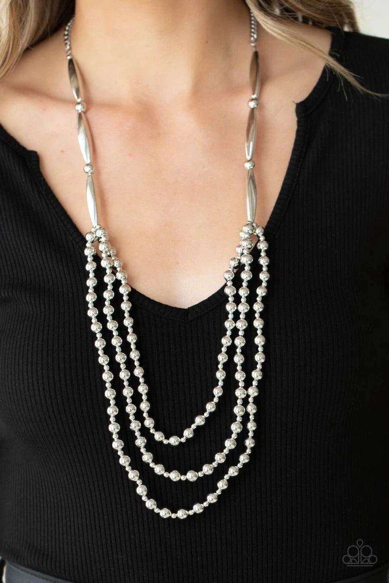 Paparazzi Accessories Beaded Beacon - Silver Brushed in a shiny shimmer, dainty and classic silver beads alternate across the chest in three gleaming layers. The tiered display attaches to oblong and classic silver beads that connect to a silver chain for