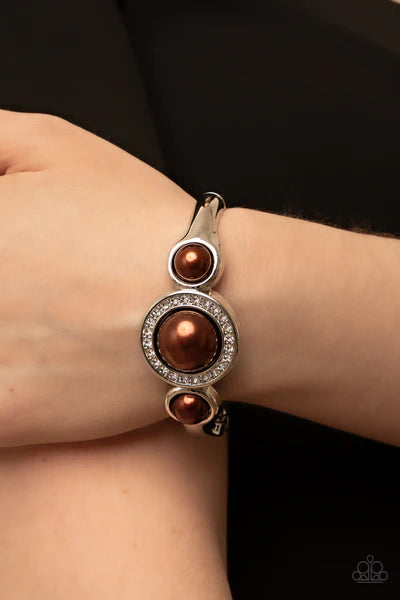 Paparazzi Accessories Debutante Daydream - Brown Encircled in a glitzy ring of glassy white rhinestones, an oversized brown pearl is flanked by a pair of bubbly brown pearl fittings atop a classic silver bangle-like bracelet for a timeless twist. Features