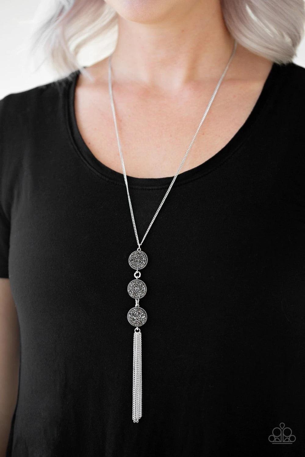 Paparazzi Accessories Triple Shimmer - Silver Sprinkled in smoky and hematite prism style rhinestones, three sparkling silver frames trickle down the chest. A shimmery silver tassel swings from the bottom of the triple stacked pendant for a glamorous fini