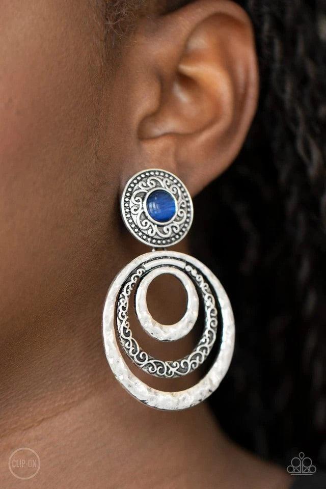 Paparazzi Accessories Bare Your Soul - Blue *Clip-On A glowing blue cat's eye stone is pressed into a studded silver frame embossed in swirling filigree. Varying in hammered and embossed textures, three silver rings ripple out from the bottom of the cat's