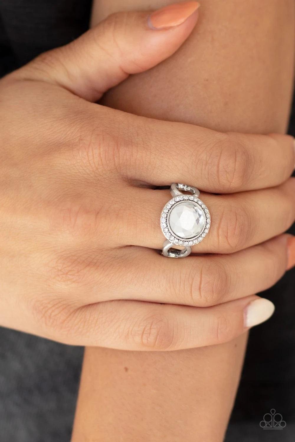 Paparazzi Accessories It’s Gonna GLOW! - White An oversized white gem is pressed into the center of a round silver frame encrusted in a ring of radiant white rhinestones for a regal flair. Features a dainty stretchy band for a flexible fit. Sold as one in