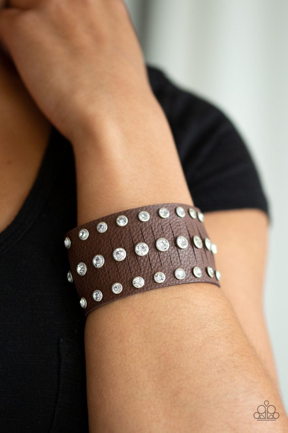 Paparazzi Accessories Now Taking The Stage - Brown Pressed into sleek silver frames, glittery white rhinestones are studded across a thick brown leather band featuring a center lined with slits for a sassy finish. Features an adjustable snap closure. Sold