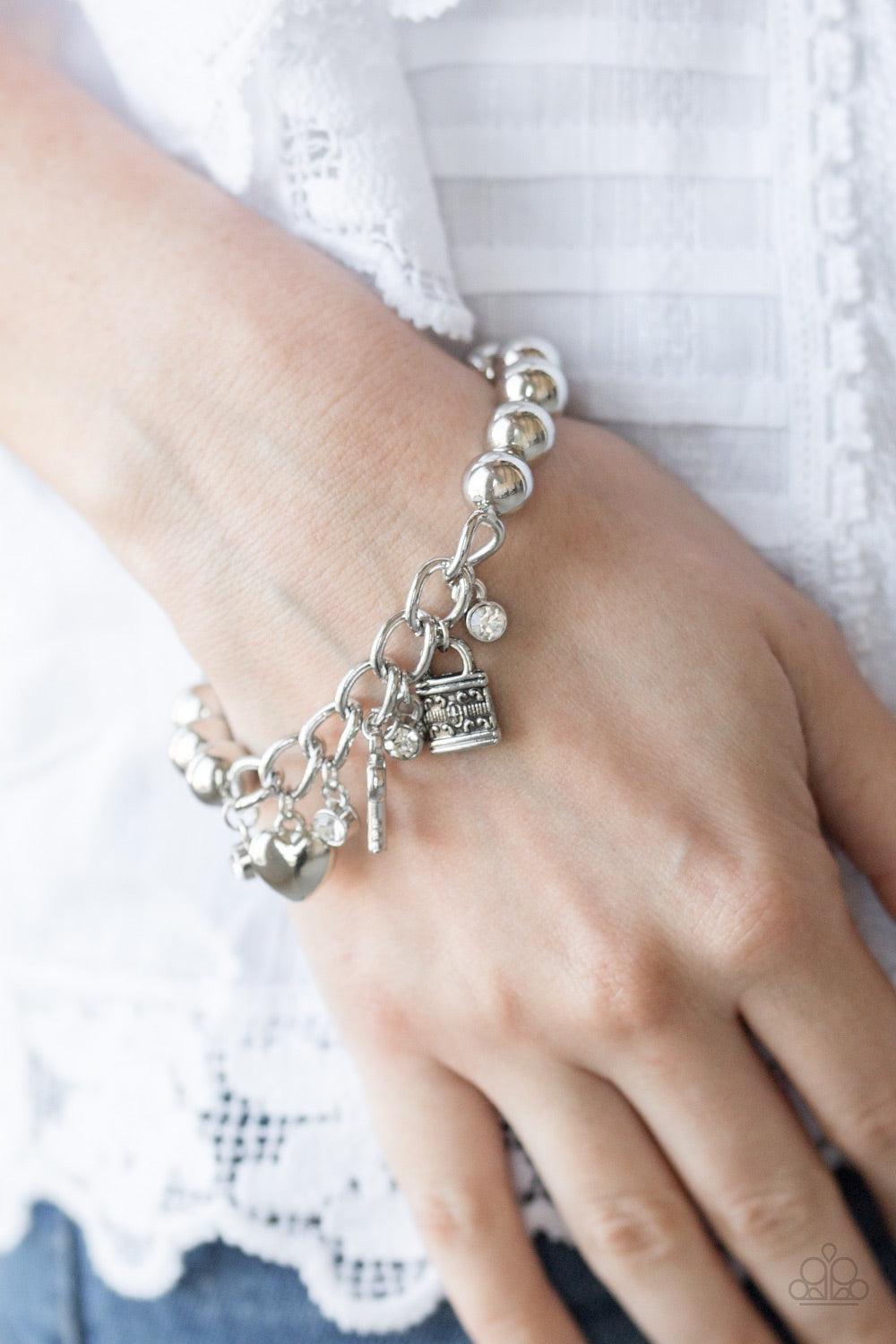 Paparazzi Accessories Feelin Flirtacious - White Attached to a section of silver chain, classic silver beads are threaded along a stretchy elastic band. Glittery white rhinestones, a silver heart, a silver key, and a silver lock swing from the wrist for a