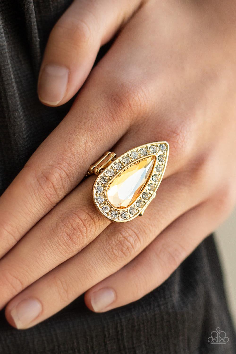Paparazzi Accessories Majestic Mayhem ~Gold An oversized golden teardrop gem is pressed in the center of a glistening gold teardrop frame radiating with glassy white rhinestones for a fierce look. Features a stretchy band for a flexible fit.