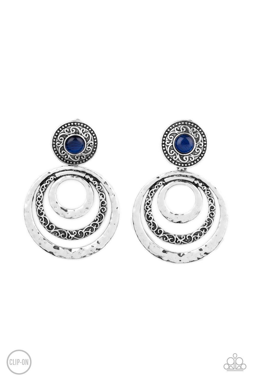 Paparazzi Accessories Bare Your Soul - Blue *Clip-On A glowing blue cat's eye stone is pressed into a studded silver frame embossed in swirling filigree. Varying in hammered and embossed textures, three silver rings ripple out from the bottom of the cat's