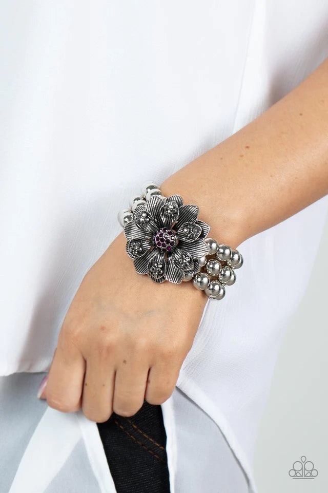 Paparazzi Accessories Botanical Bravado - Purple A daring oversized silver flower is composed of petals lined in antiqued silver and dotted with smoky hematite rhinestones. A sphere of dainty purple rhinestones creates the center of the flower as it sits