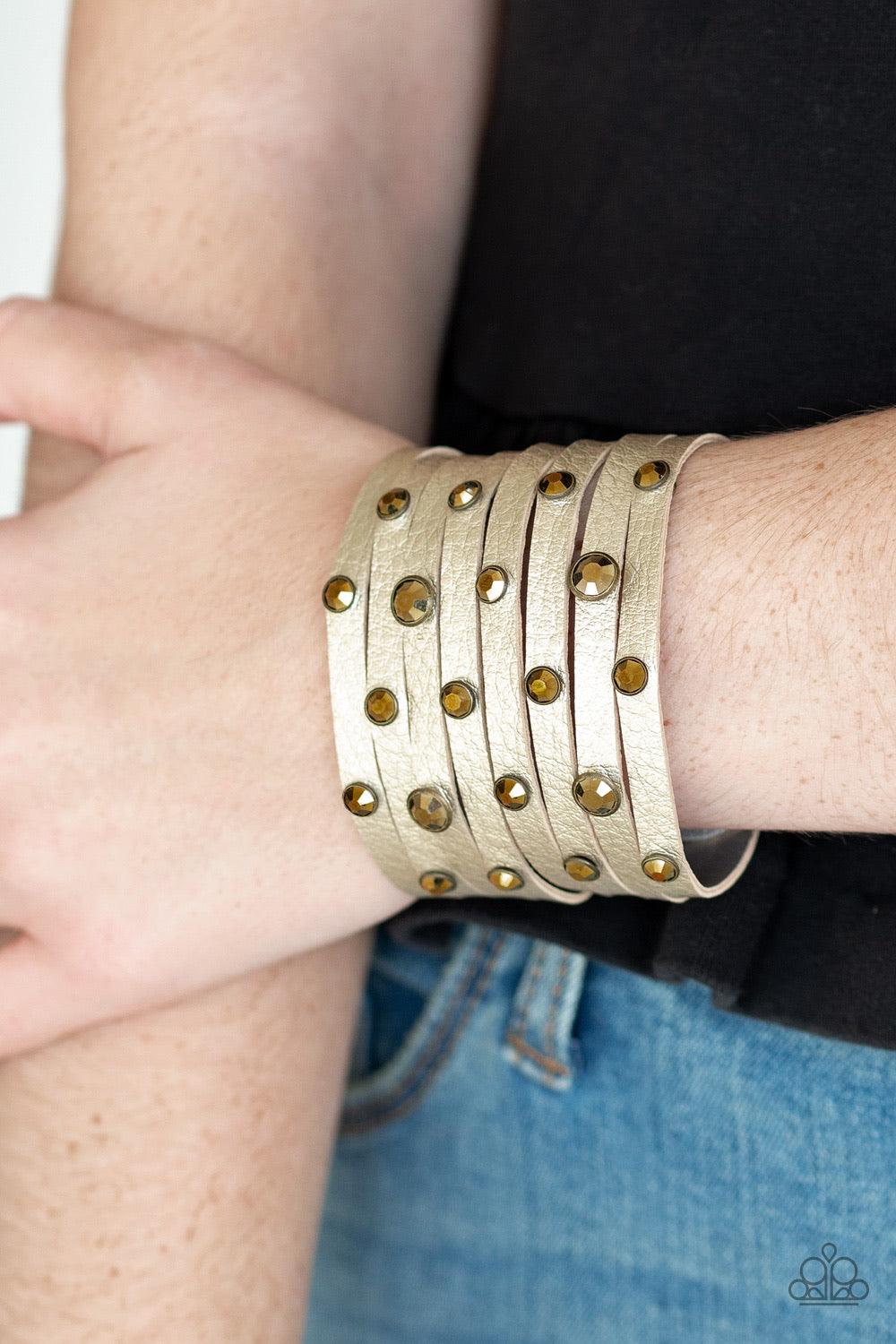 Paparazzi Accessories Go-Getter Glamorous - Brass Brushed in a metallic shimmer, a thick leather band has been spliced into eight strips across the wrist. Encased in sleek brass fittings, glittery aurum rhinestones are sprinkled across the center for a sa