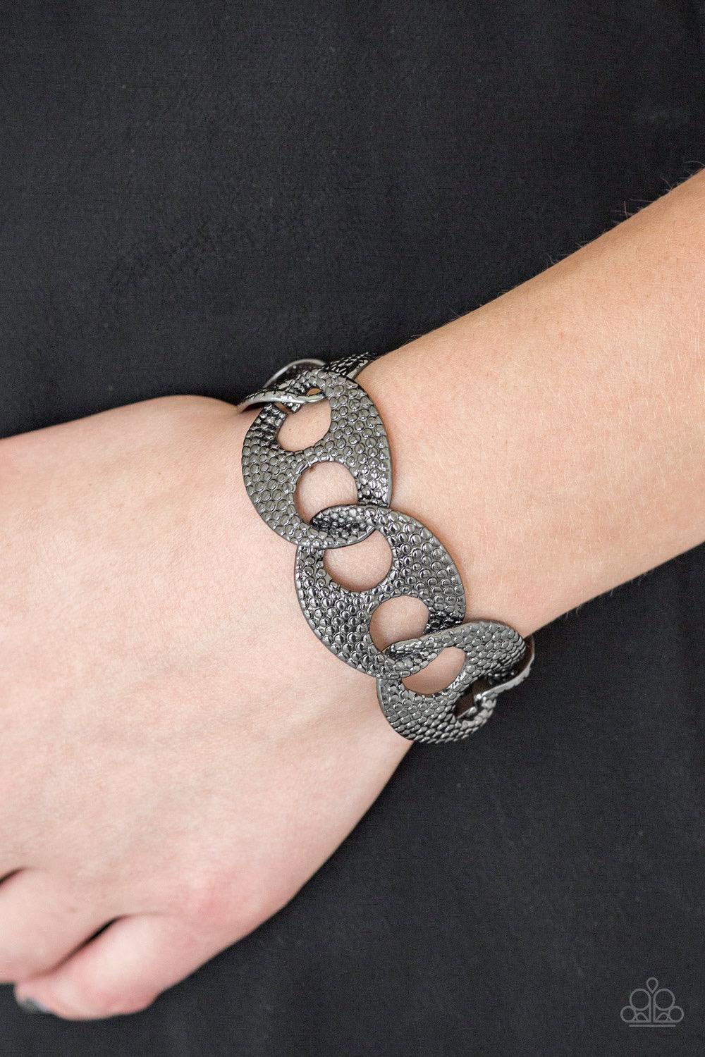 Paparazzi Accessories Casual Connoisseur - Black Embossed in shimmery circular patterns, asymmetrical gunmetal frames link across the wrist for a casually industrial look. Features an adjustable clasp closure. Jewelry