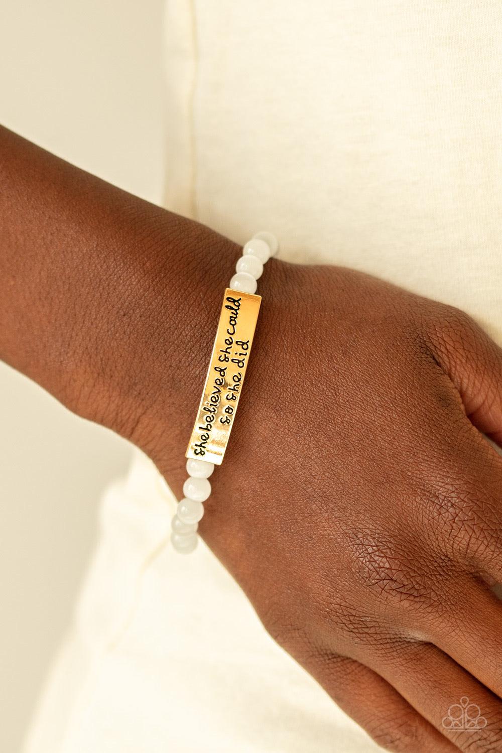 Paparazzi Accessories So She Did - Gold A collection of dainty white cat’s eye stone beads and a gold frame stamped in the inspirational phrase, “She believed she could, so she did”, are threaded along a stretchy band around the wrist for a whimsical fash