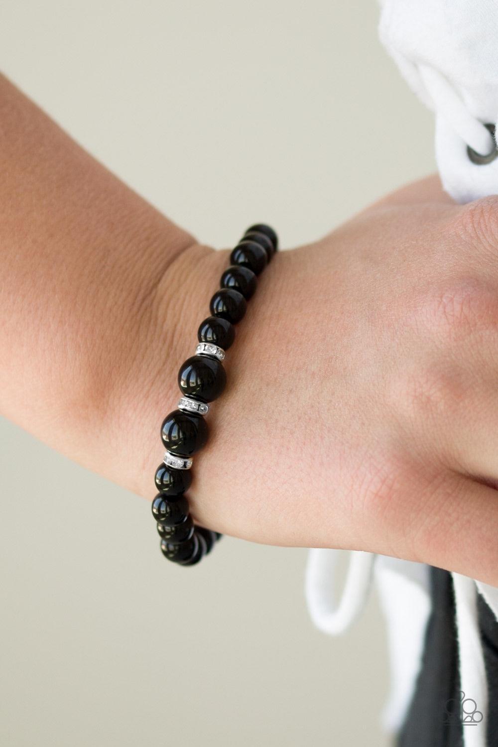 Paparazzi Accessories Radiantly Royal - Black Gradually increasing in size near the center, shiny black beads are threaded along a stretchy elastic band. White rhinestone encrusted rings are sprinkled along the center for a timeless finish. Jewelry